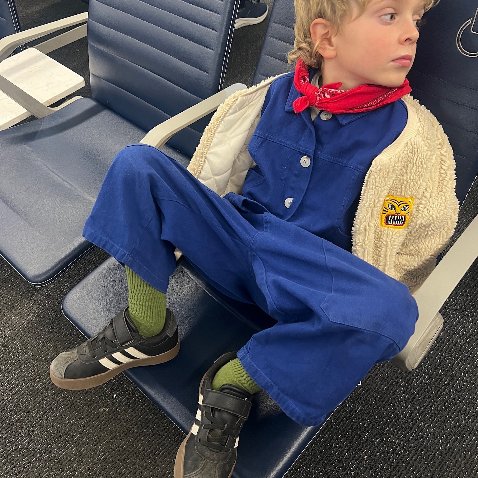 Child sitting on an airplane seat wearing a blue jumpsuit with a red bandana.