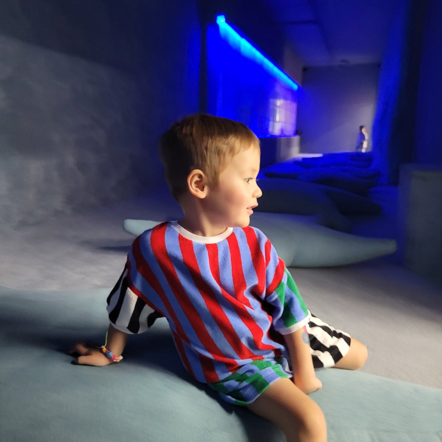 Child in a colorful striped shirt sitting on a bed in a dimly lit room with blue lighting.
