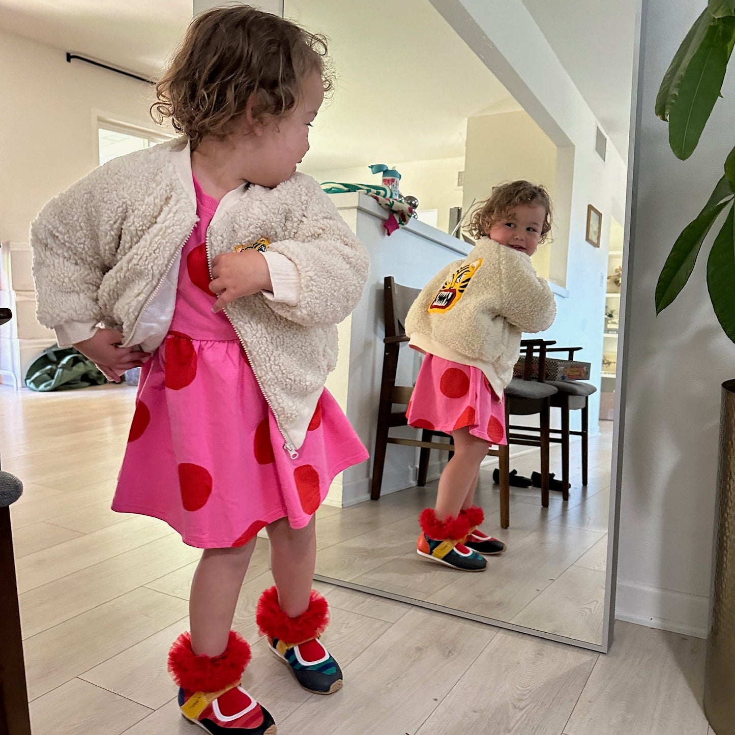 Child in a pink dress with red hearts standing in front of a mirror.