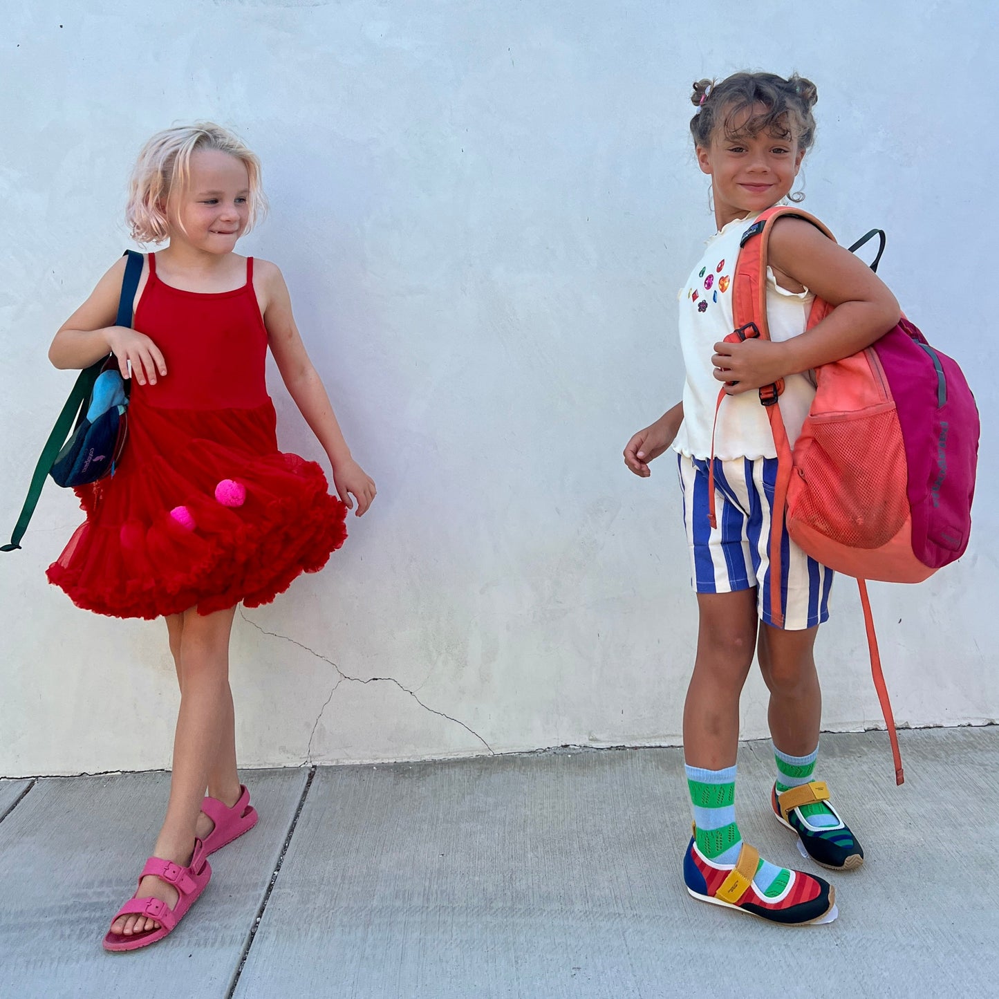 Two children standing against a white wall, one in a red dress with a blue bag and the other in colorful striped shorts with a pink bag.