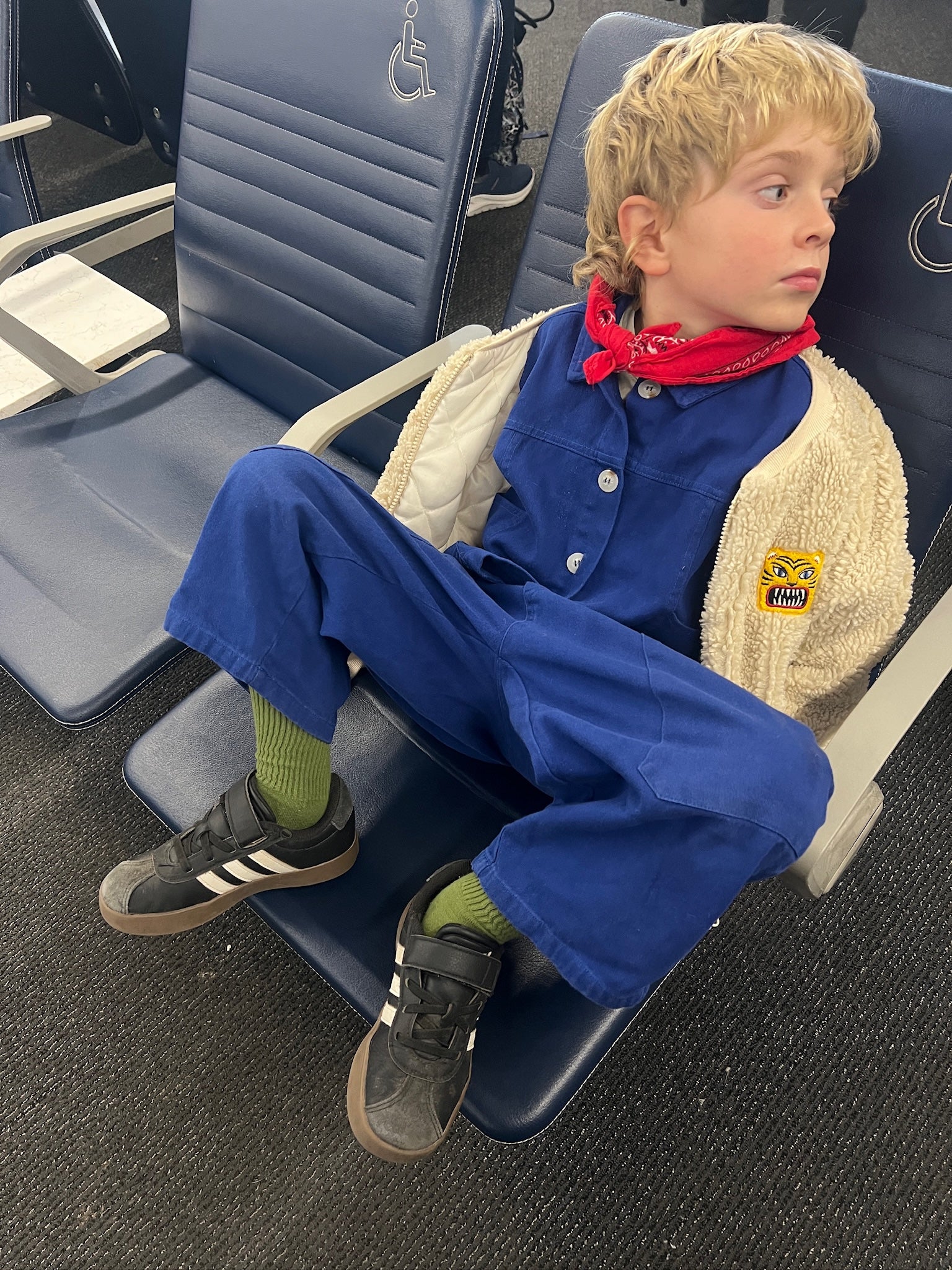 Child sitting on an airplane seat wearing a blue jumpsuit with a red bandana.