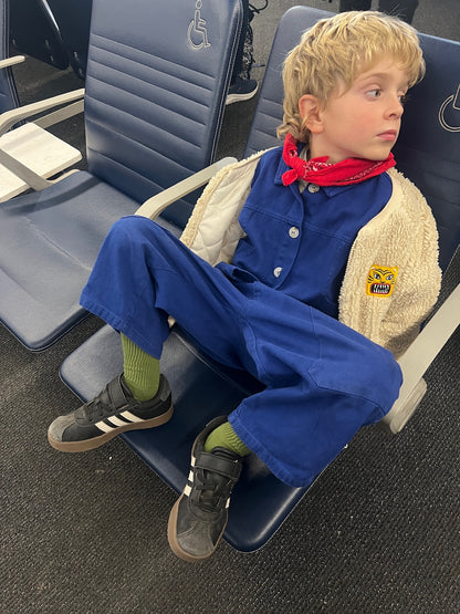 Child sitting on an airplane seat wearing a blue jumpsuit with a red bandana.