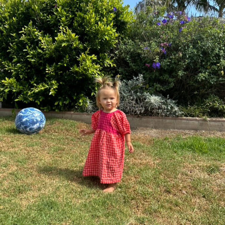 Child in a red dress standing on grass with bushes and flowers in the background