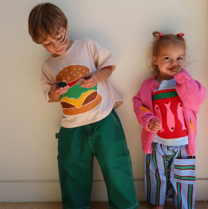 Two children standing against a white wall, one wearing a shirt with a burger design and the other in a pink sweater with animal prints.
