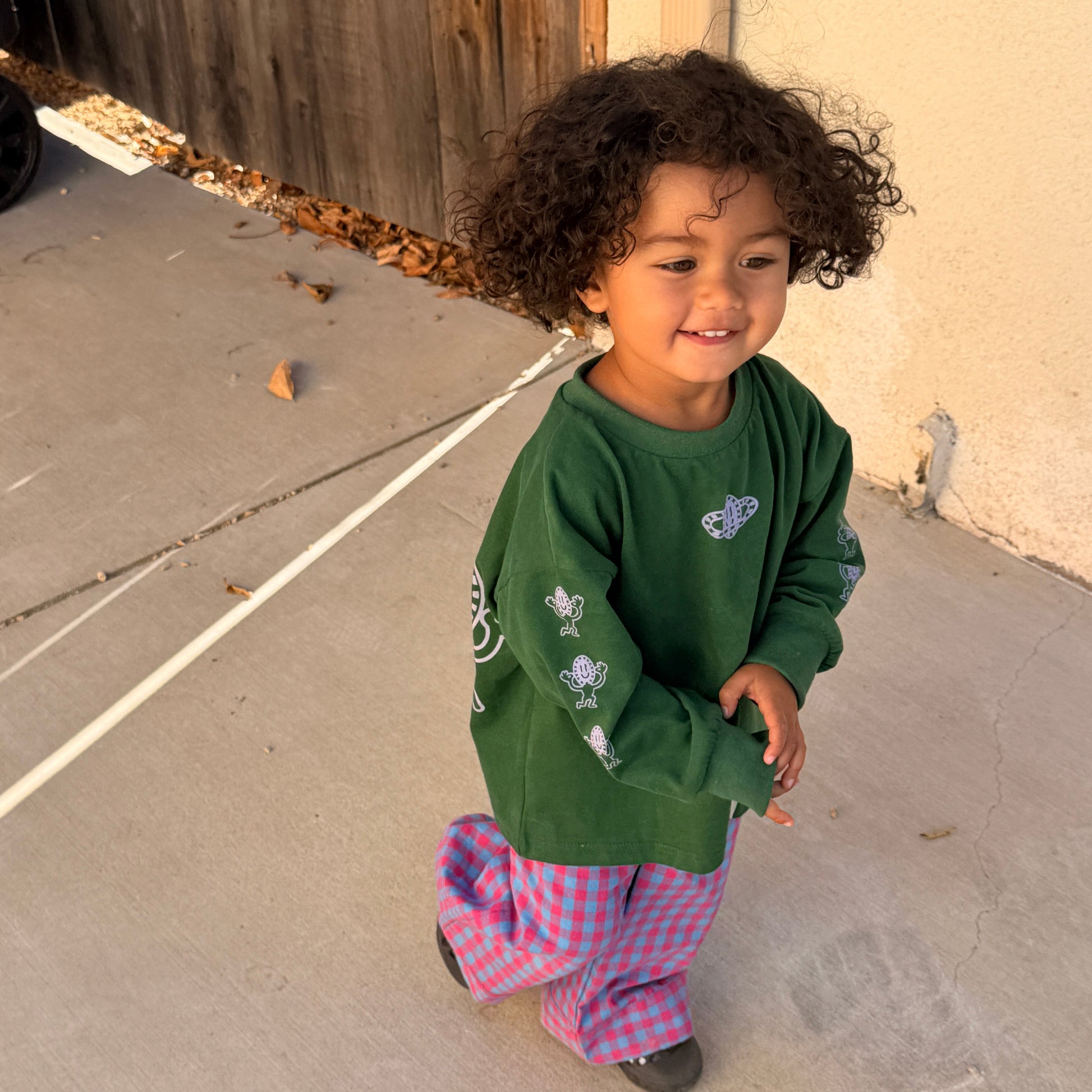 Child in a green hoodie and plaid pants standing on a sidewalk.
